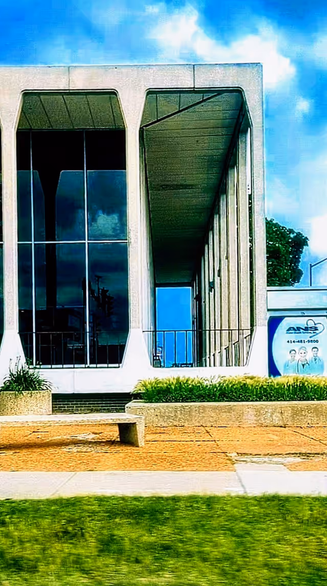 Modern building front with large glass windows, entrance, and landscaped walkway.