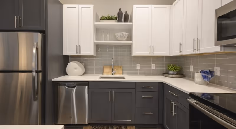 Modern kitchen with stainless steel refrigerator, dishwasher, and oven. The cabinetry is two-toned with dark lower cabinets and white upper cabinets. There is a small sink with a faucet in the center, open shelves with decorative items, and a gray tiled backsplash.