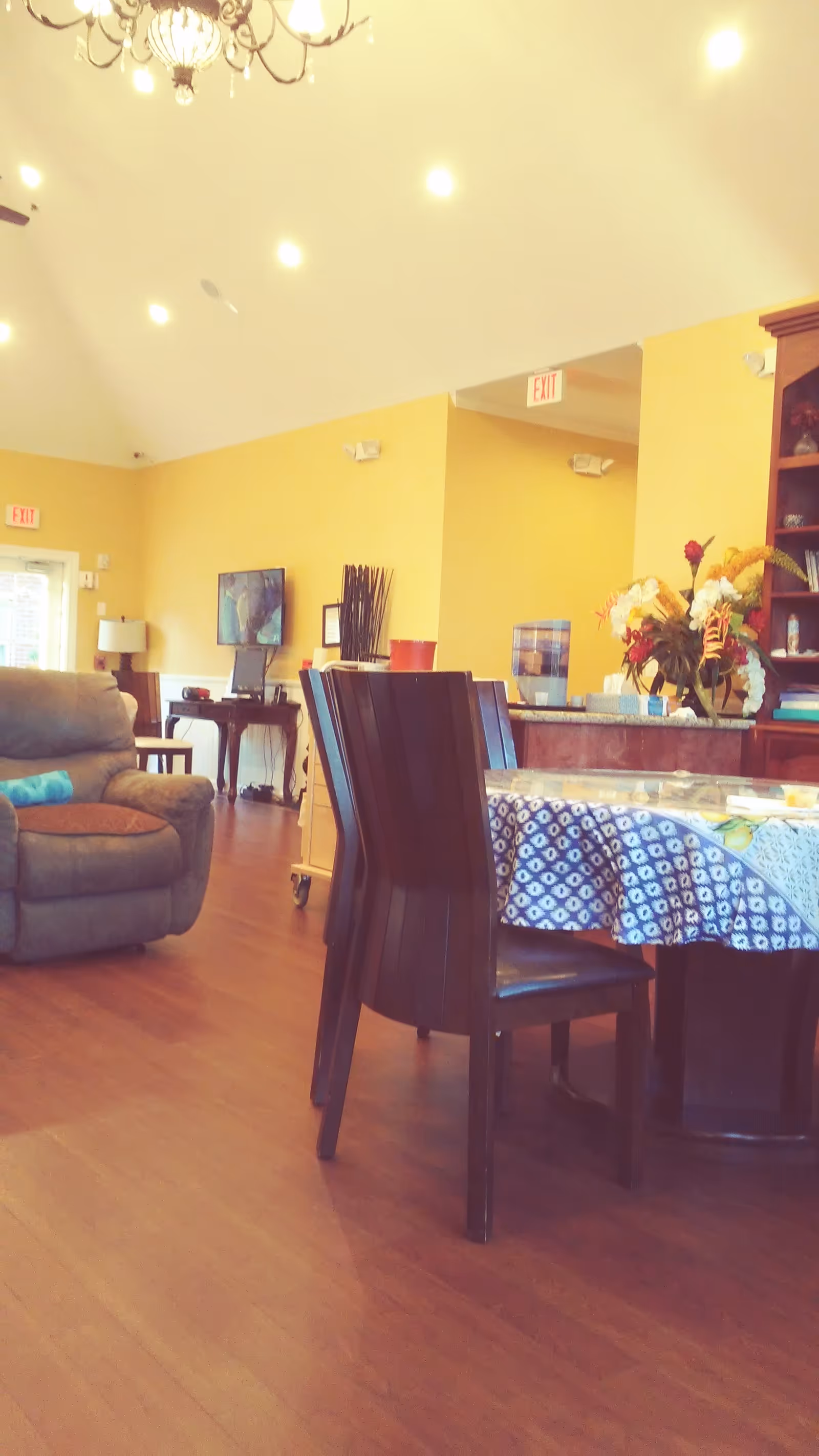 Interior view of a yellow-walled senior living common area with couch, dining table, chandelier.