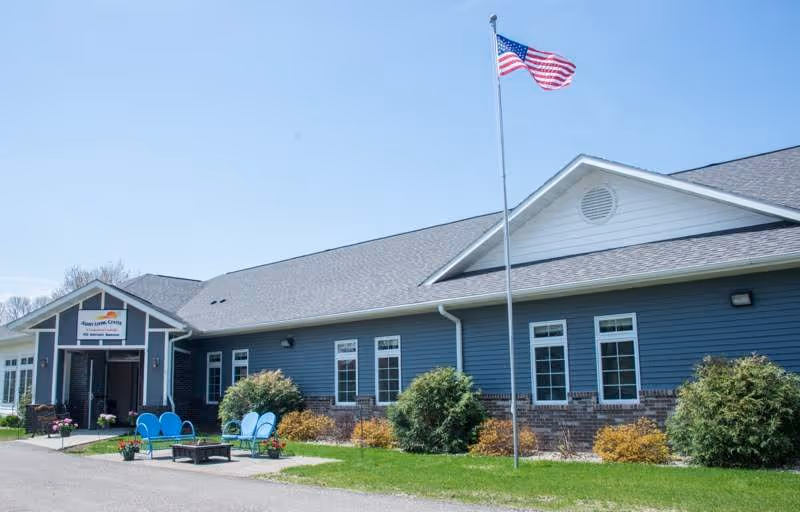 Exterior view of Ashby Living Center, a single-story building with blue siding and brick accents. There is a flagpole with an American flag in front, along with outdoor seating including blue chairs and a fire pit on a paved area. The sky is clear and blue.