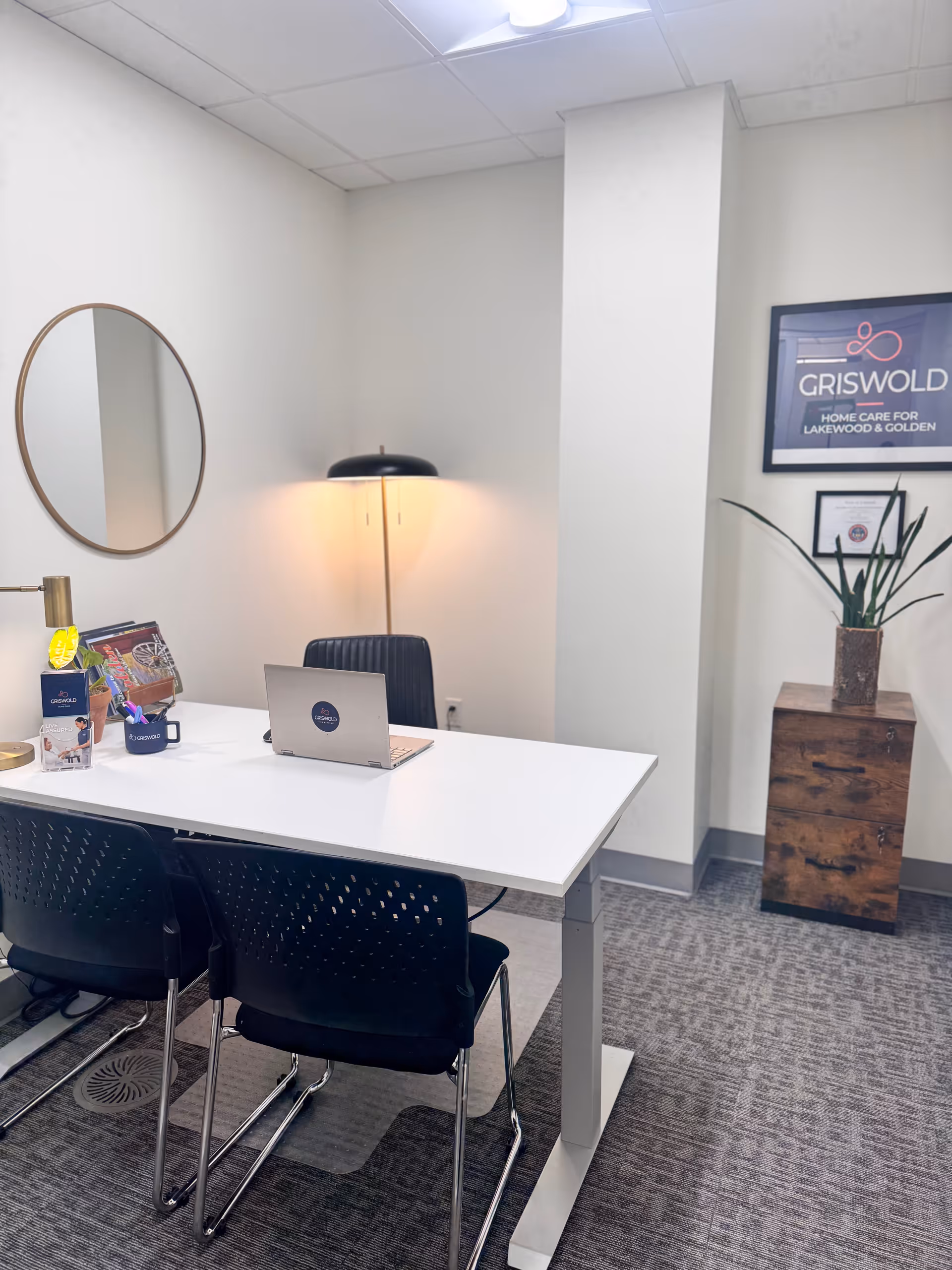 White office desk with laptop, chairs, mirror, lamp, and Griswold sign in a reception area.