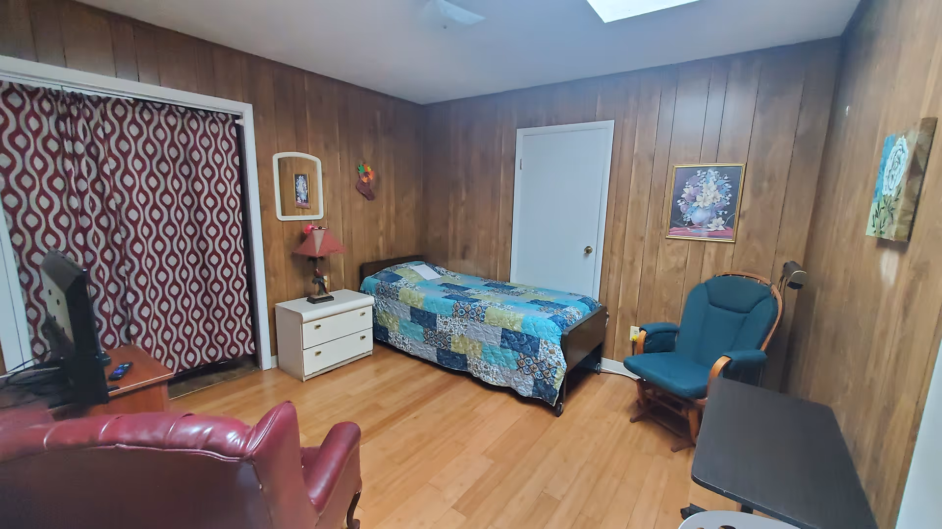 Wood-paneled senior living bedroom featuring a single bed with a patchwork quilt, dresser, chair, and TV.