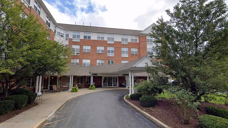 Front entrance of a multi-story brick senior living building with a covered porte-cochère, driveway, and landscaped trees and shrubs.