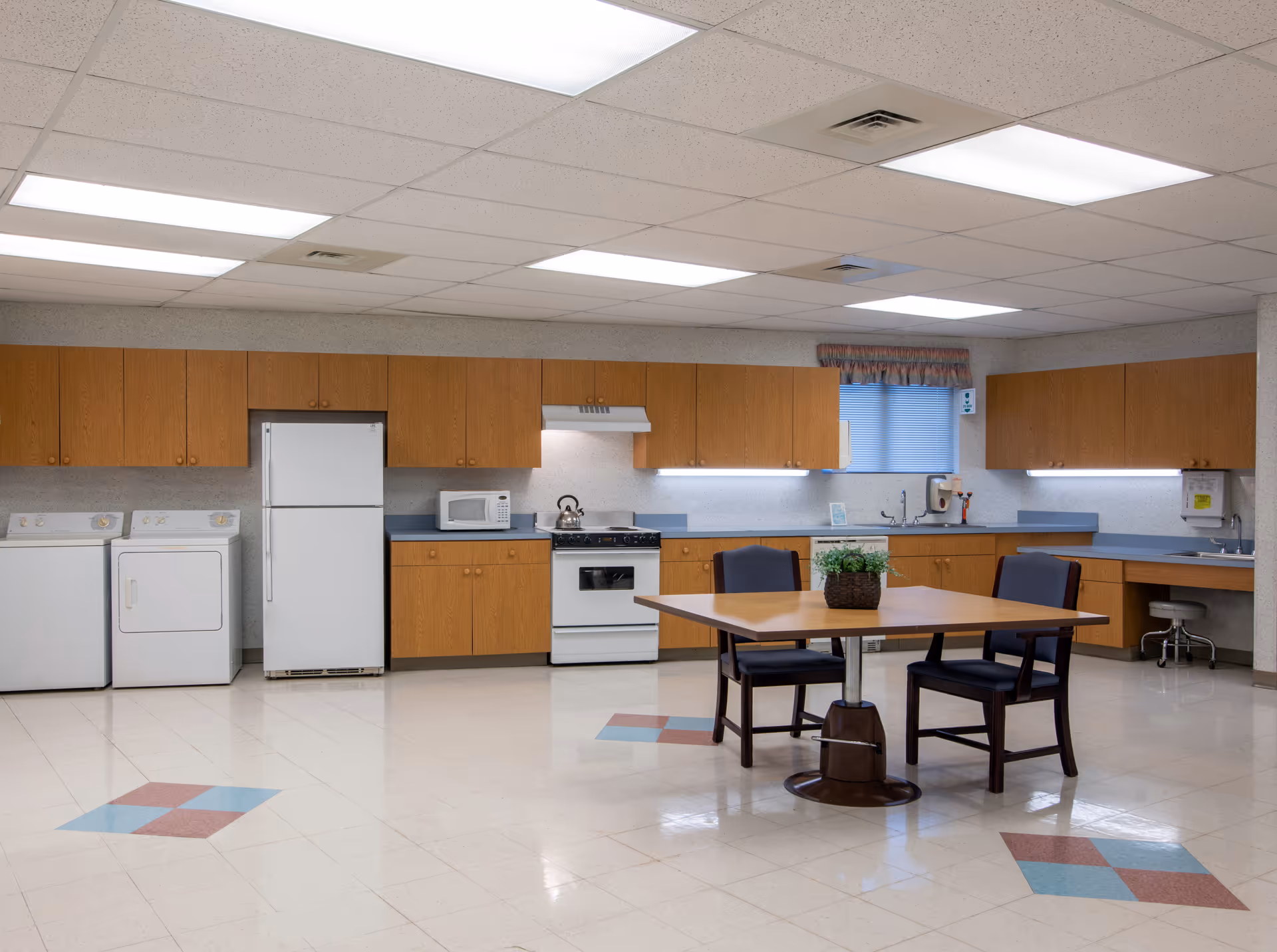 A spacious kitchen area in a senior living facility with wooden cabinets, a white refrigerator, stove, microwave, and laundry machines. There is a wooden table with two chairs and a small plant on the table. The floor has a tiled pattern with blue and red diamond shapes, and the ceiling has fluorescent lighting panels.