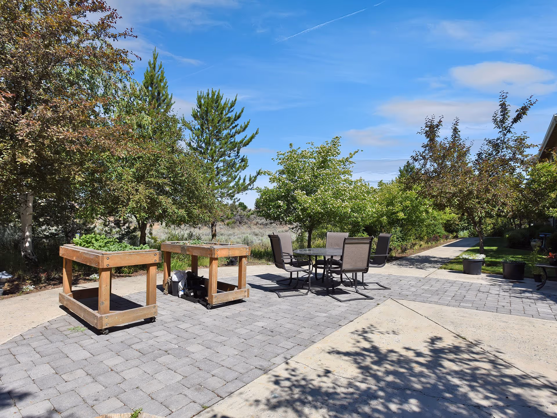 Outdoor patio area with a paved stone surface featuring a round glass table surrounded by four chairs. Two wooden planter boxes with plants are positioned on the left side. The background includes various green trees and shrubs under a blue sky with some clouds.