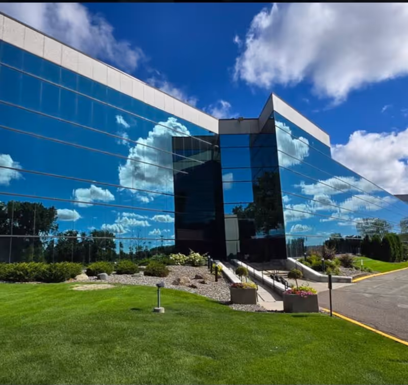Modern glass-front building with reflecting blue sky, landscaped lawn, and accessible entrance ramp.