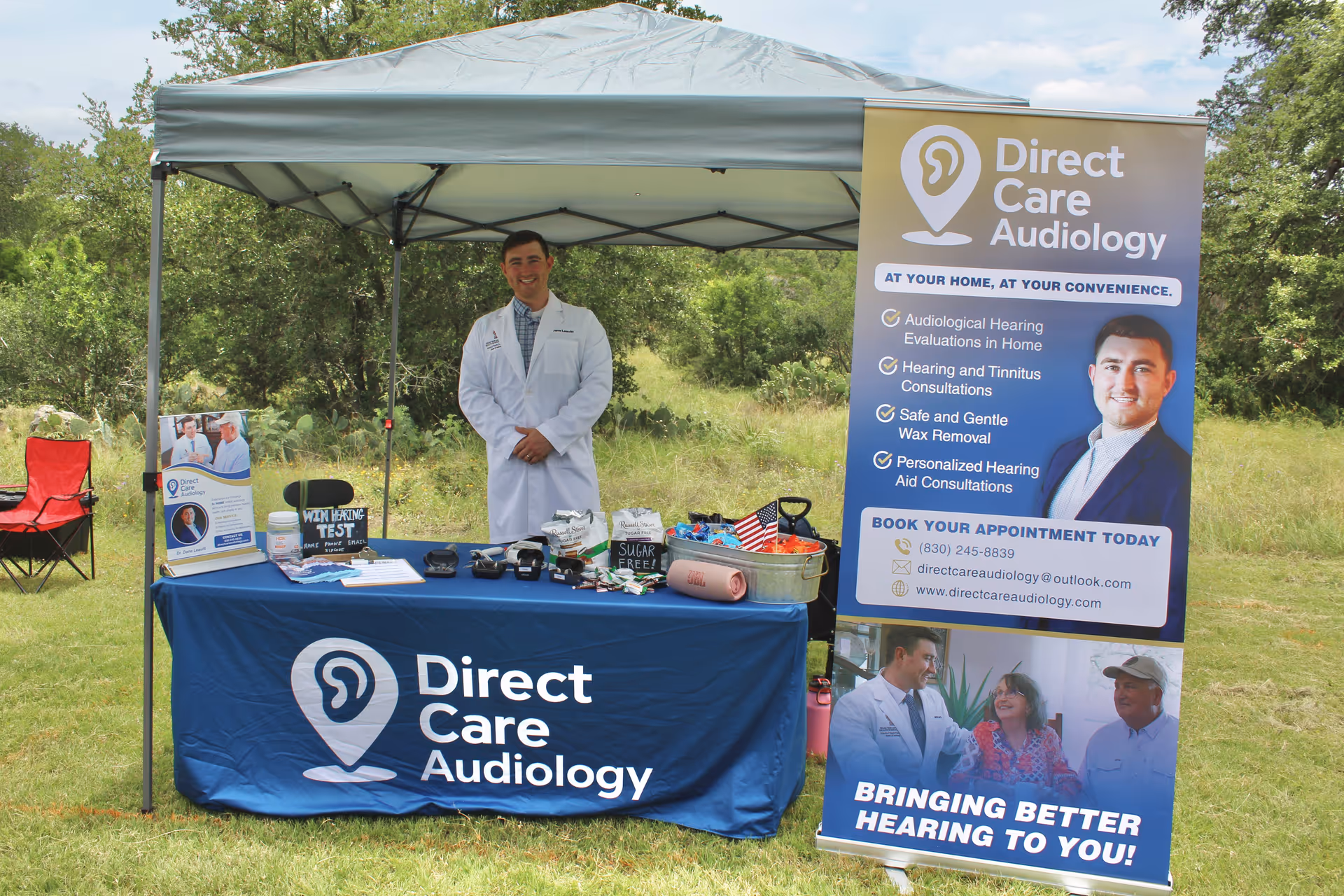 An outdoor Direct Care Audiology booth with a tent, table, banners, and a doctor.