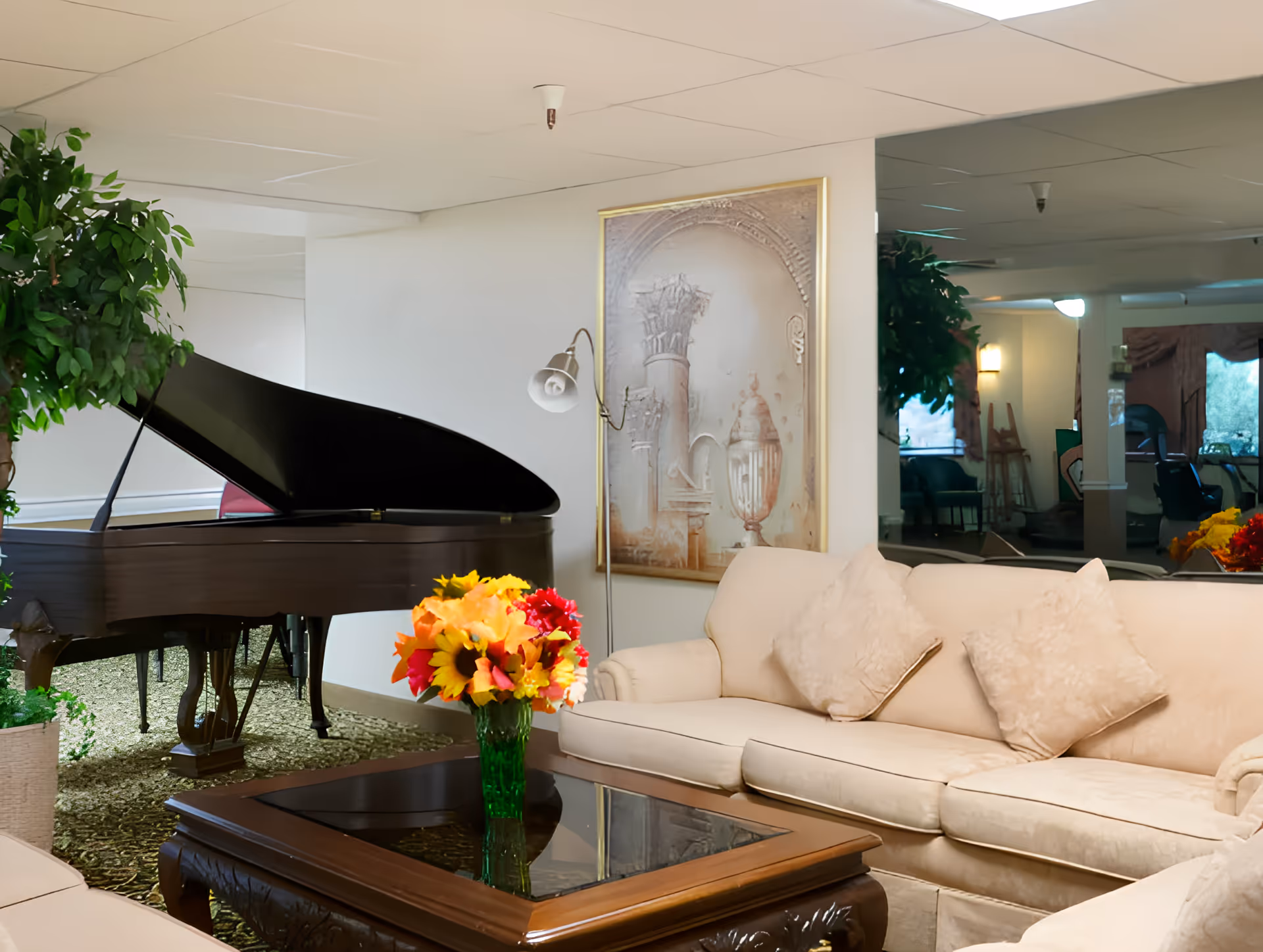 Lounge area with a grand piano, beige sofas, a wooden coffee table holding a vase of colorful flowers, artwork and potted plants.