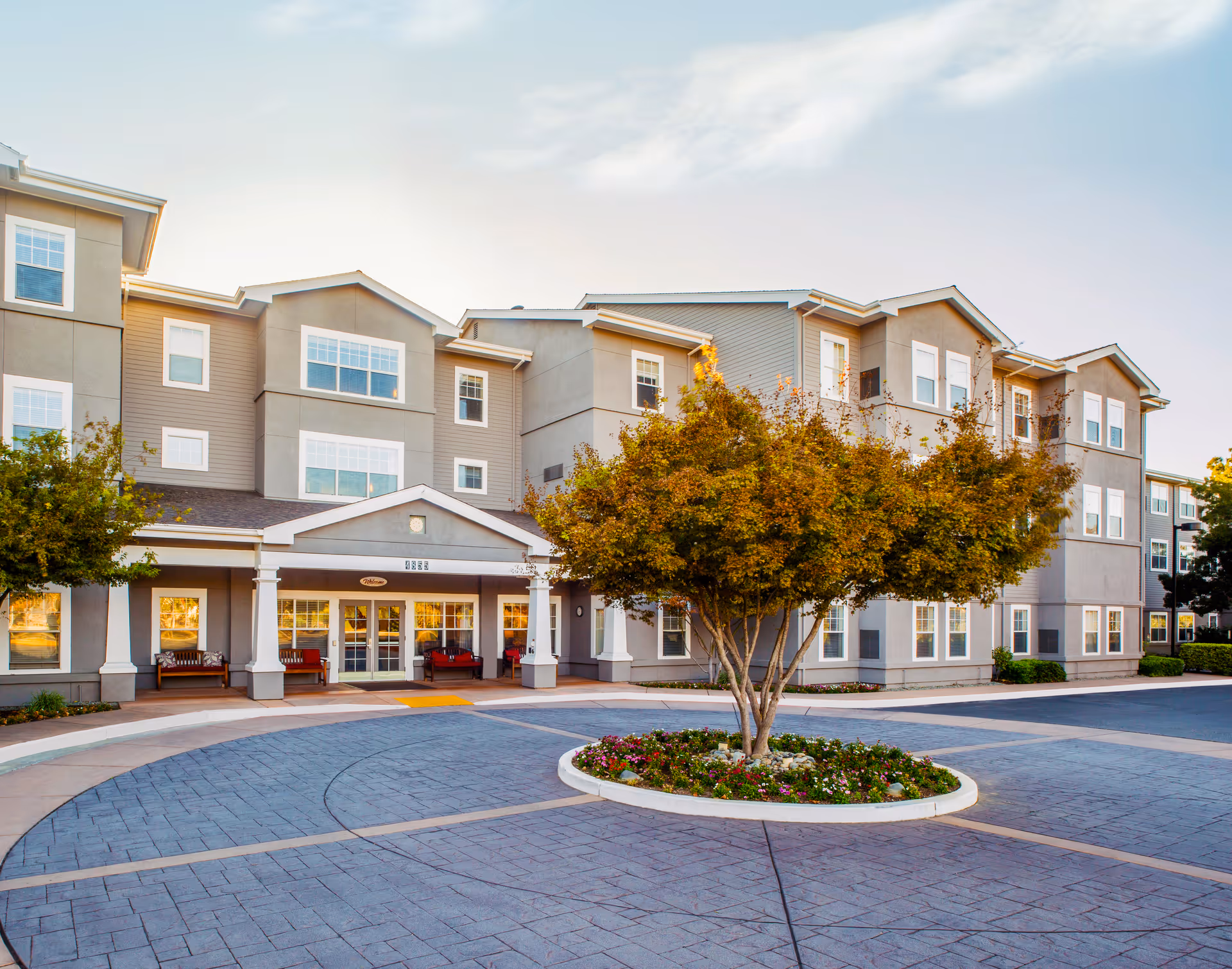 Front entrance of a multi-story senior living building with a circular driveway and a tree and flower bed in the center.