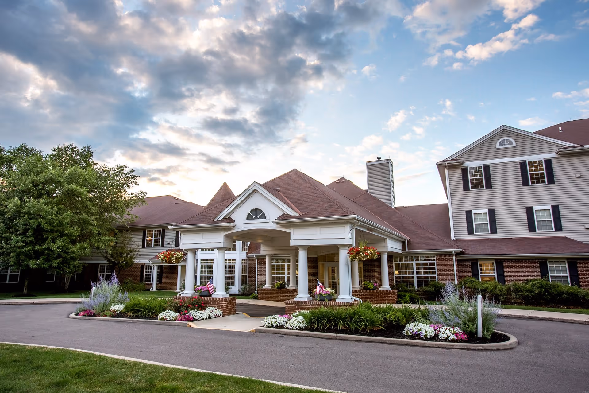 Exterior view of a senior living facility building with a covered entrance supported by white columns, landscaped flower beds, and a partly cloudy sky in the background.
