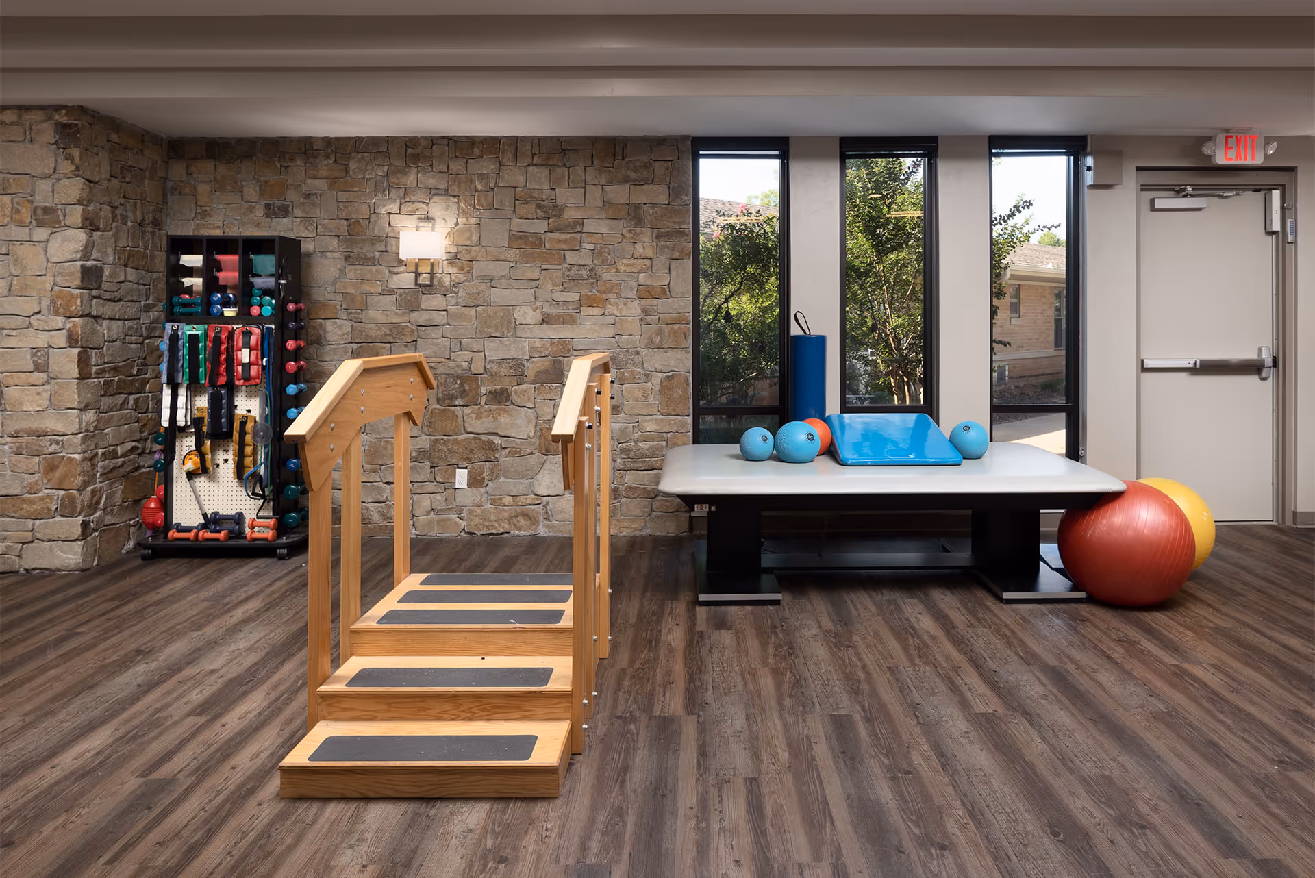 A physical therapy room with wooden parallel bars for walking exercises, a padded therapy table with exercise balls and mats, a rack of various exercise equipment, stone accent wall, large windows showing greenery outside, and a door with an exit sign above it.