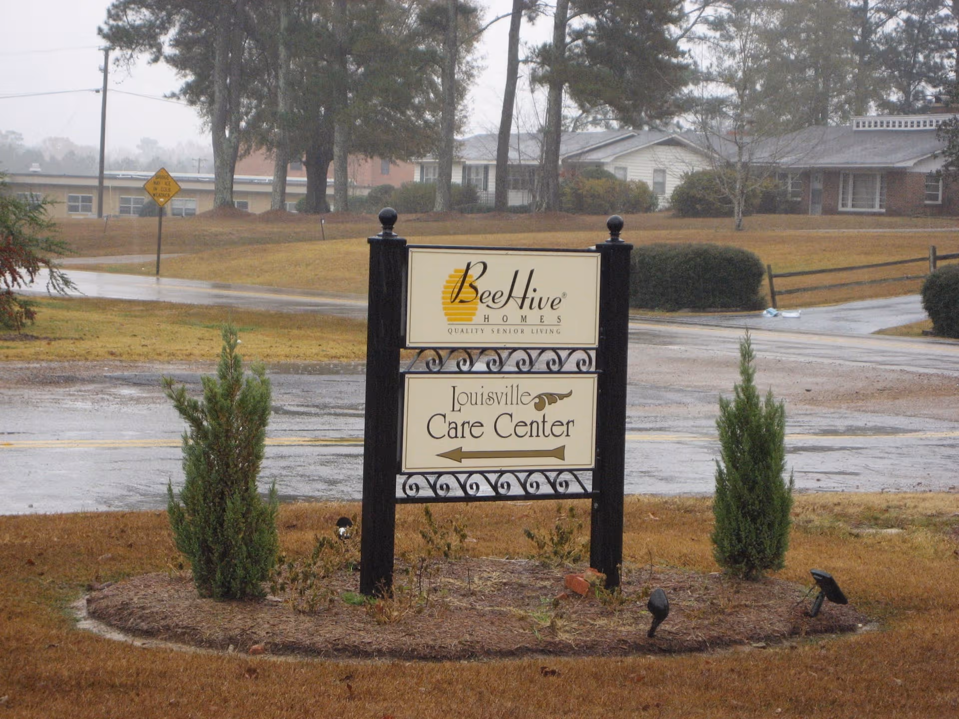 Outdoor BeeHive Homes Louisville Care Center sign in a rainy, grassy area.