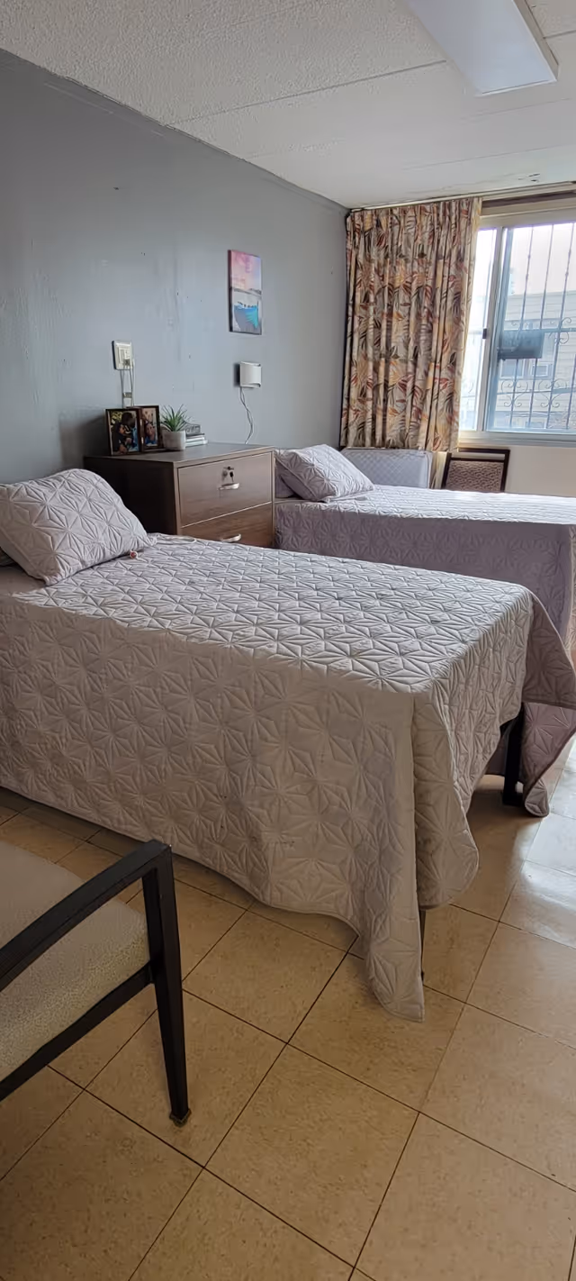 A simple bedroom with two single beds covered in light-colored patterned bedspreads. Between the beds is a wooden nightstand with framed photos and a small plant. A window with patterned curtains allows natural light into the room. The floor is tiled and there is a chair partially visible in the foreground.