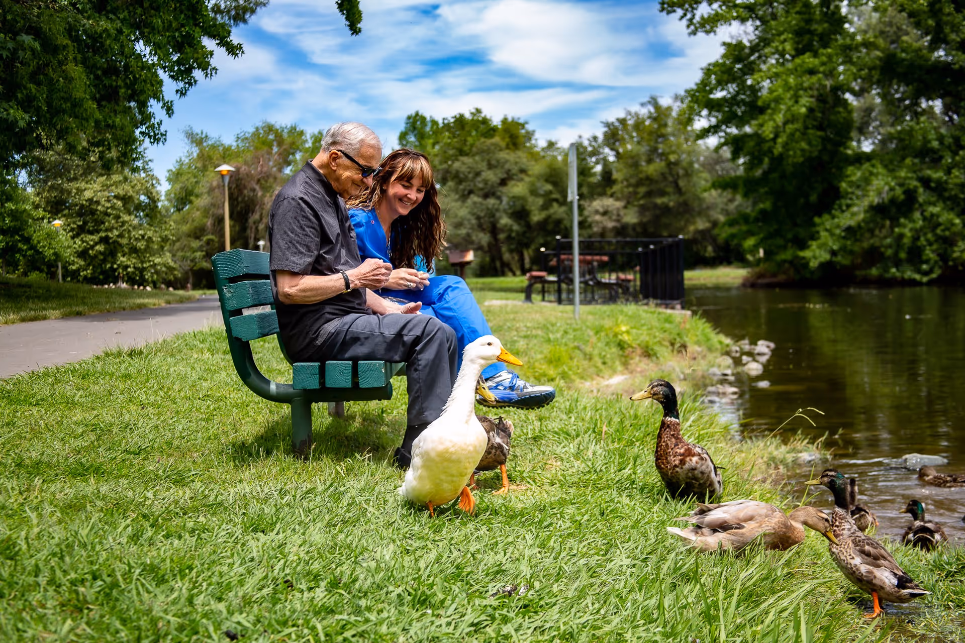 An older man and a woman sit on a green bench by a lake, feeding ducks.