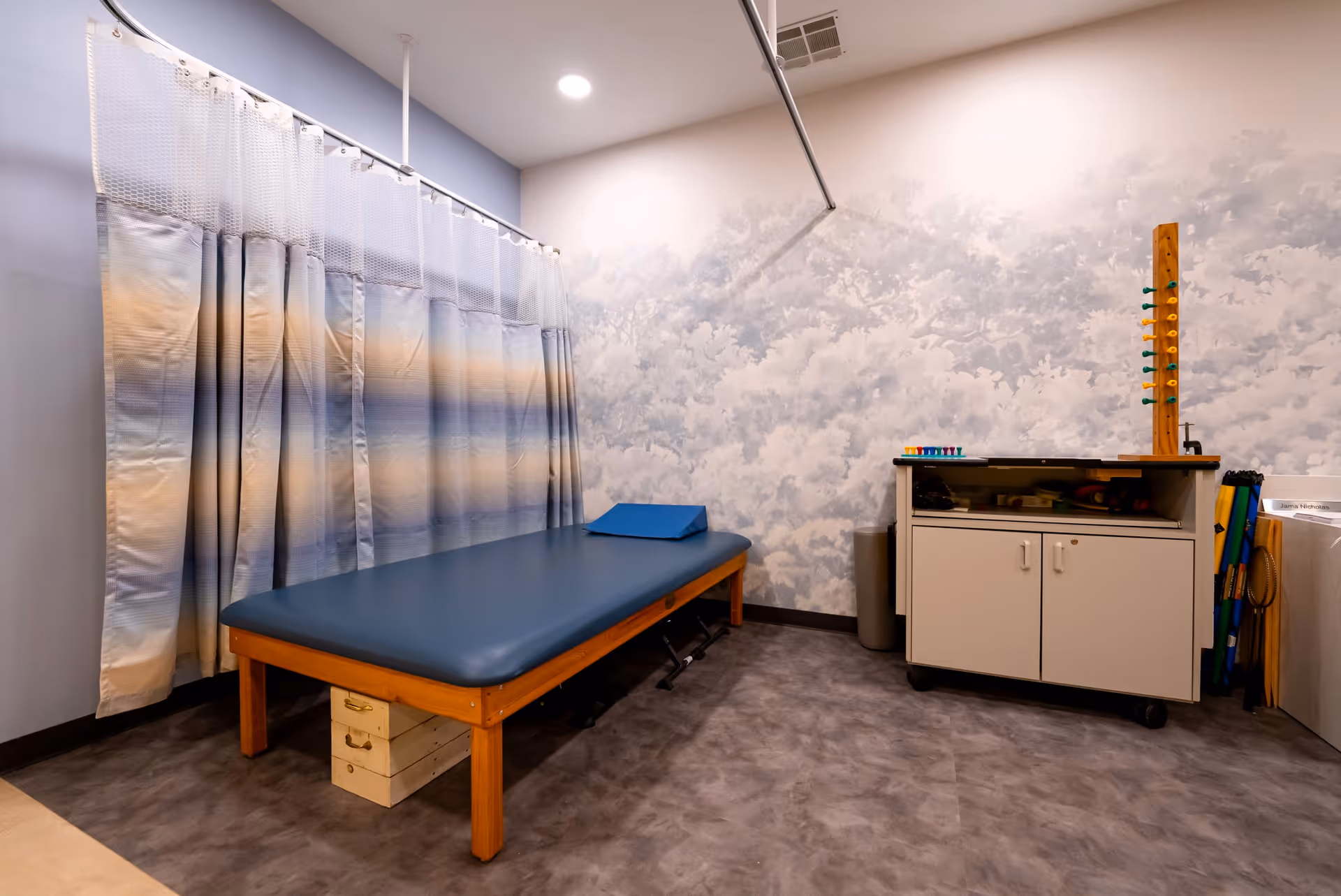 A treatment room with a blue padded therapy table, patterned privacy curtain, and a storage cabinet against a cloud-patterned wall.