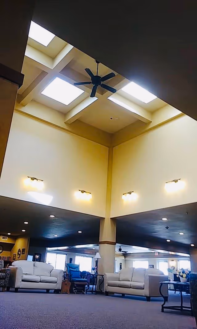 Spacious living room area with high ceilings featuring skylights and a ceiling fan. The room is furnished with white sofas, a blue recliner, and side tables. Warm wall lights illuminate the space, and large windows in the background allow natural light to enter.