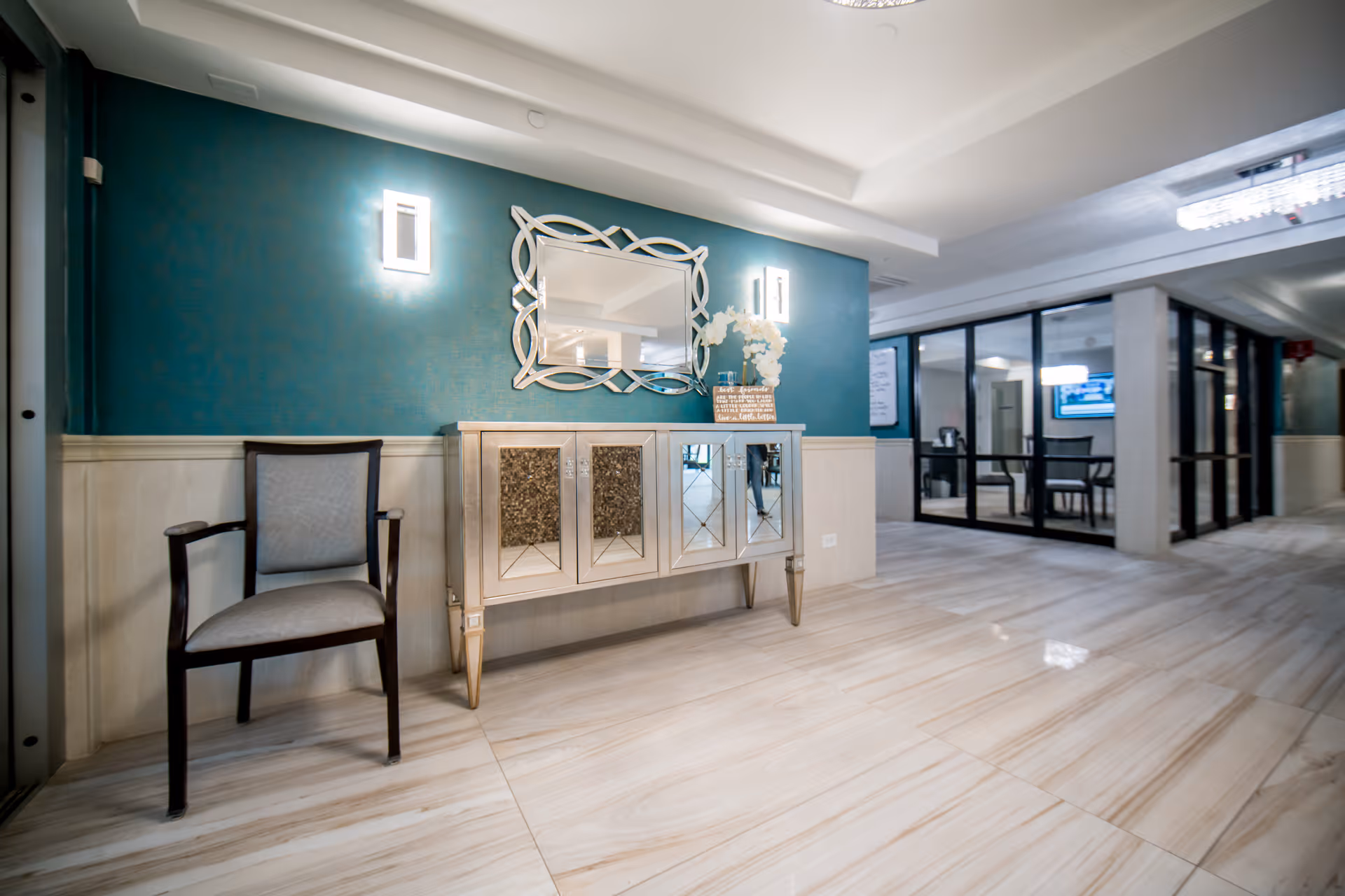 A well-lit interior hallway with a teal accent wall featuring a decorative mirror and two wall sconces. Below the mirror is a mirrored cabinet with a floral arrangement and a small sign on top. To the left of the cabinet is a single cushioned chair with wooden arms. The floor is tiled with light-colored marble, and the hallway extends to a glass-walled room with chairs and a television visible inside.