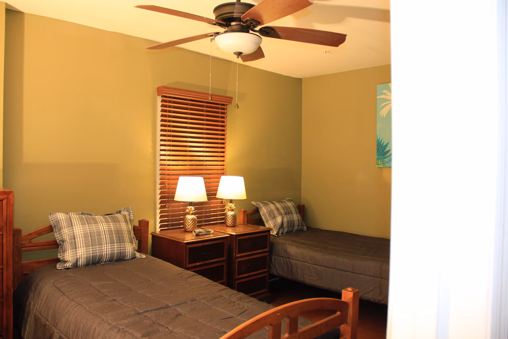 A bedroom with two single wooden beds, each with a plaid pillow and dark bedding. Between the beds is a wooden nightstand with two pineapple-shaped lamps and a phone. The walls are painted olive green, and there is a window with wooden blinds behind the nightstand. A ceiling fan with wooden blades is mounted on the ceiling.