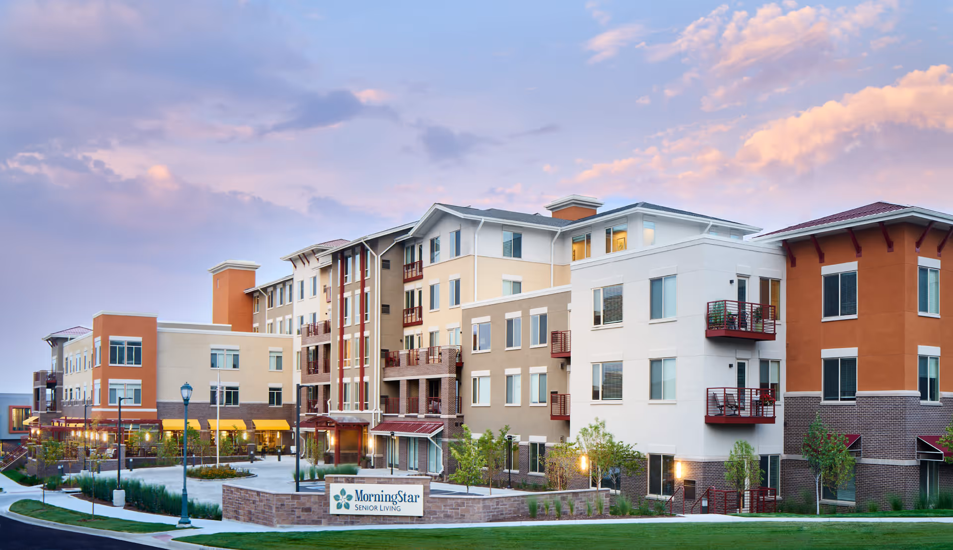 Exterior view of a modern multi-story senior living facility named MorningStar Senior Living at RidgeGate, with a well-maintained lawn, trees, and a clear sky with clouds at sunset.
