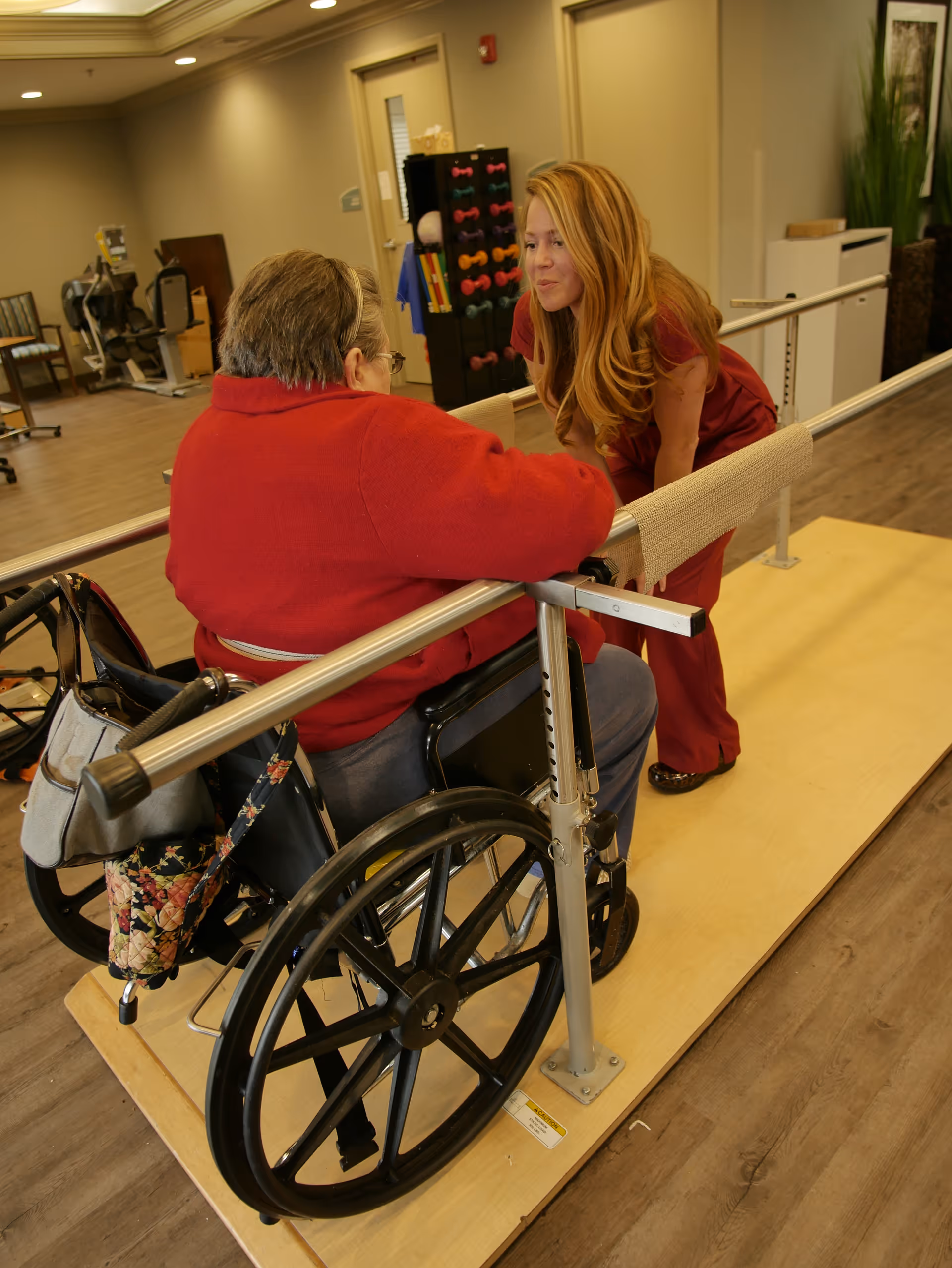 A healthcare worker in red scrubs is assisting an elderly person in a wheelchair as they practice walking with parallel bars in a rehabilitation center. The room has exercise equipment and wooden flooring.