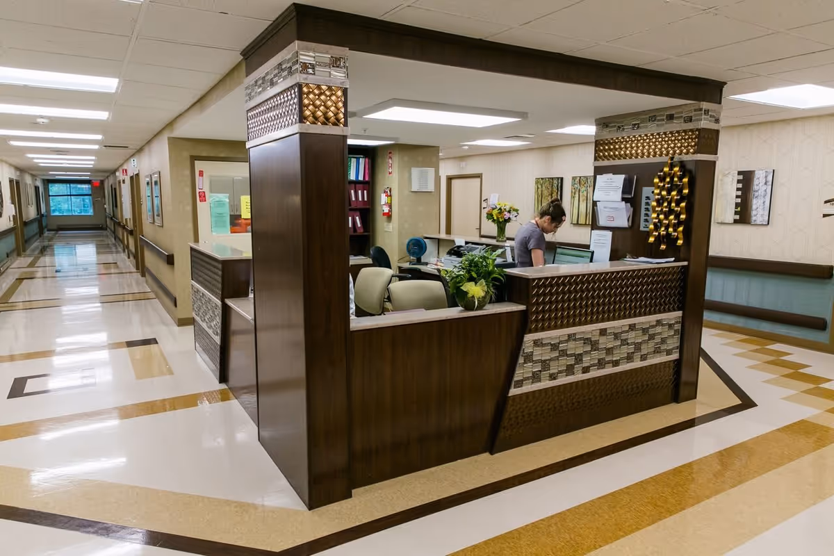A nurses' station/reception desk in a brightly lit rehabilitation center hallway with a staff member working at a computer.