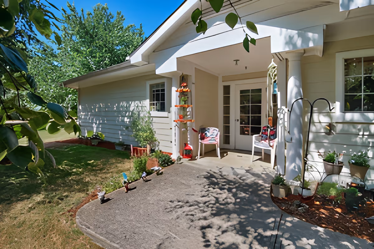 Covered front porch of a light-colored single-story building with chairs, potted plants, and a concrete walkway.