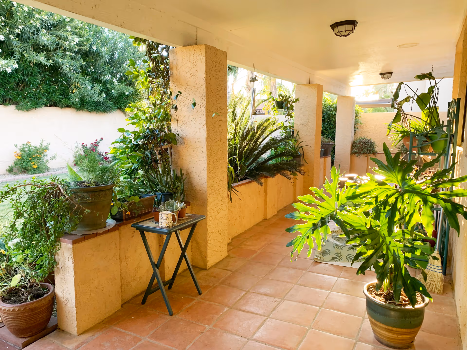 A covered outdoor patio area with terracotta tiled flooring and beige stucco walls. The space is decorated with various potted plants and greenery along the edges and on a small folding table, with sunlight illuminating the area.