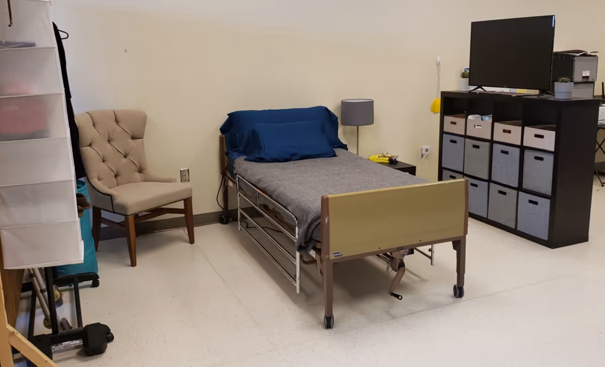 A furnished patient bedroom with a bed, chair, lamp, and storage cubes.