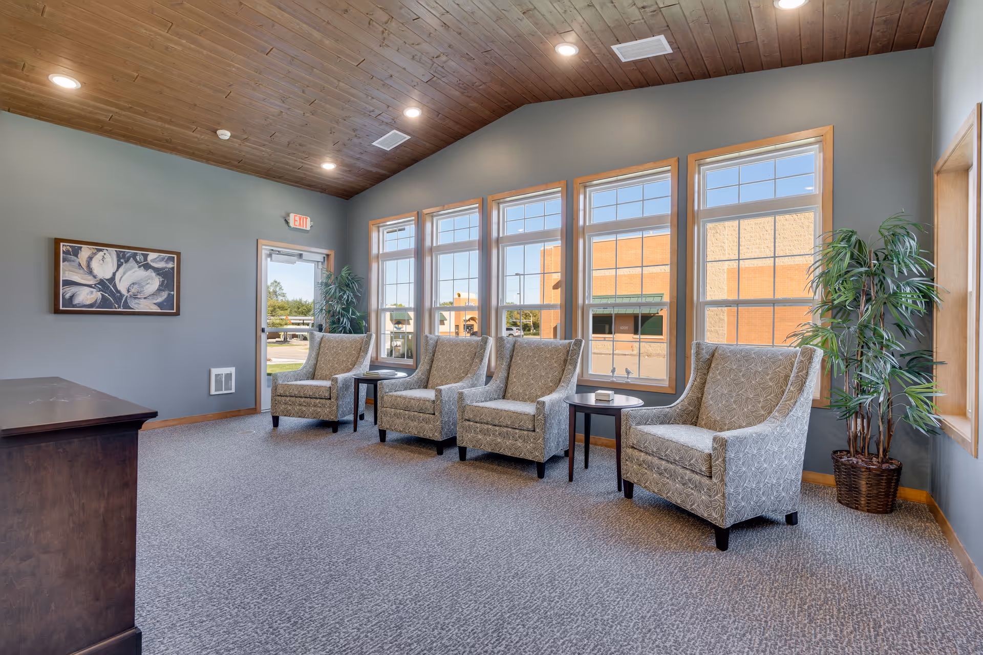 A bright sitting area with four upholstered armchairs, side tables, large windows, potted plants, and a wood-paneled ceiling.