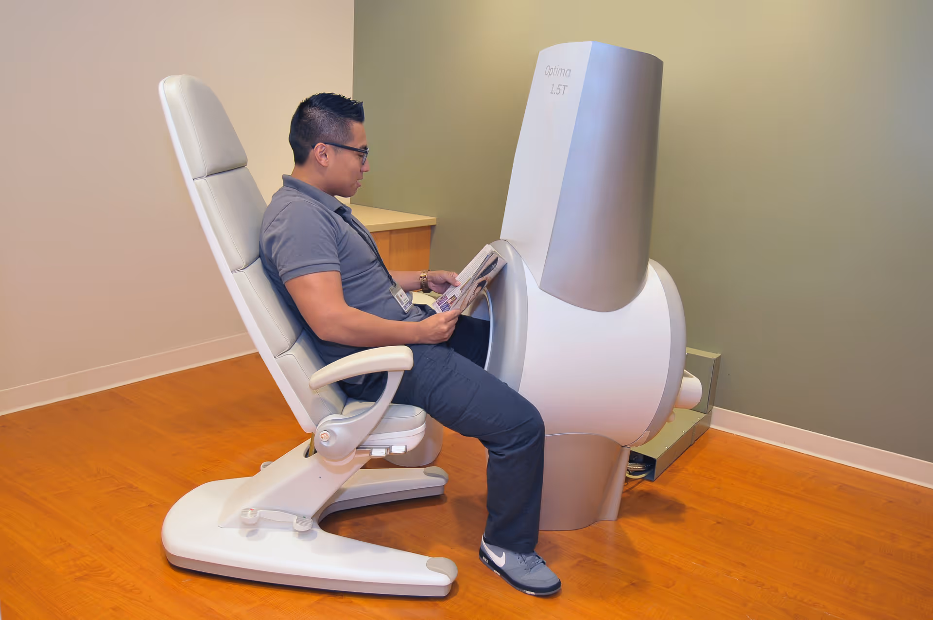 A man sits in a white reclining chair beside a gray medical device labeled Optima 157.
