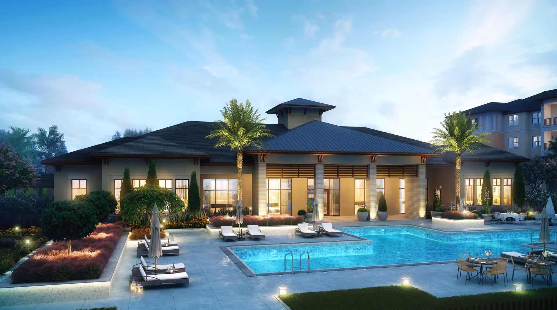 Outdoor swimming pool and patio with lounge chairs and palm trees in front of a well-lit community building at dusk.