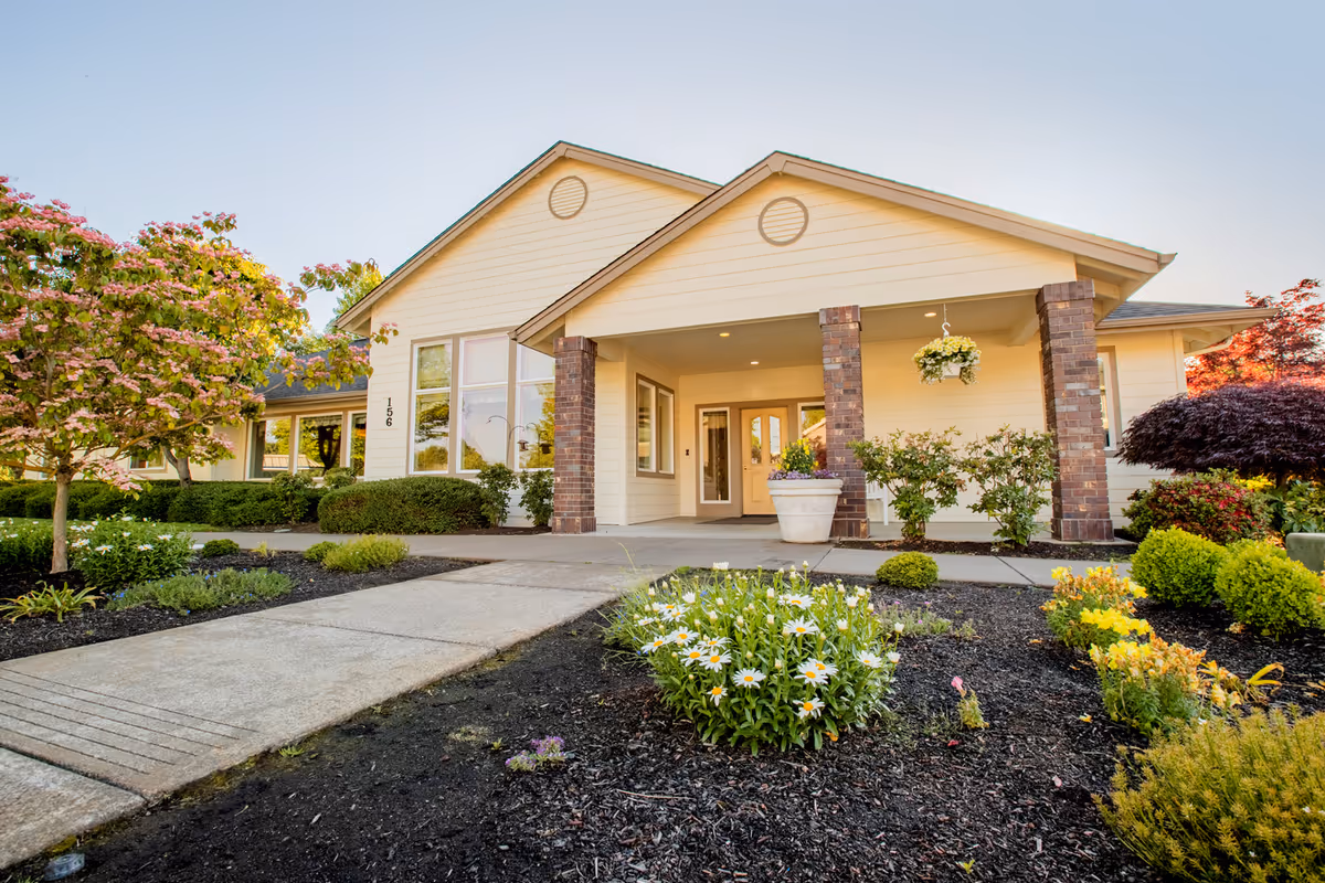 Front entrance of a single-story senior living building with a covered porch, brick columns, walkway, and landscaped flower beds.