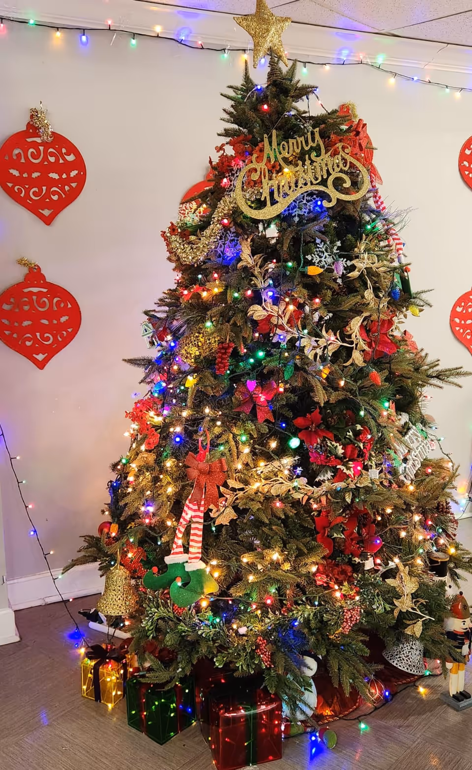 A decorated Christmas tree with multicolored lights, red and gold ornaments, a gold star topper, and a 'Merry Christmas' sign. There are wrapped gift boxes and a nutcracker figure at the base of the tree. The background shows a white wall with red holiday decorations and string lights along the ceiling.