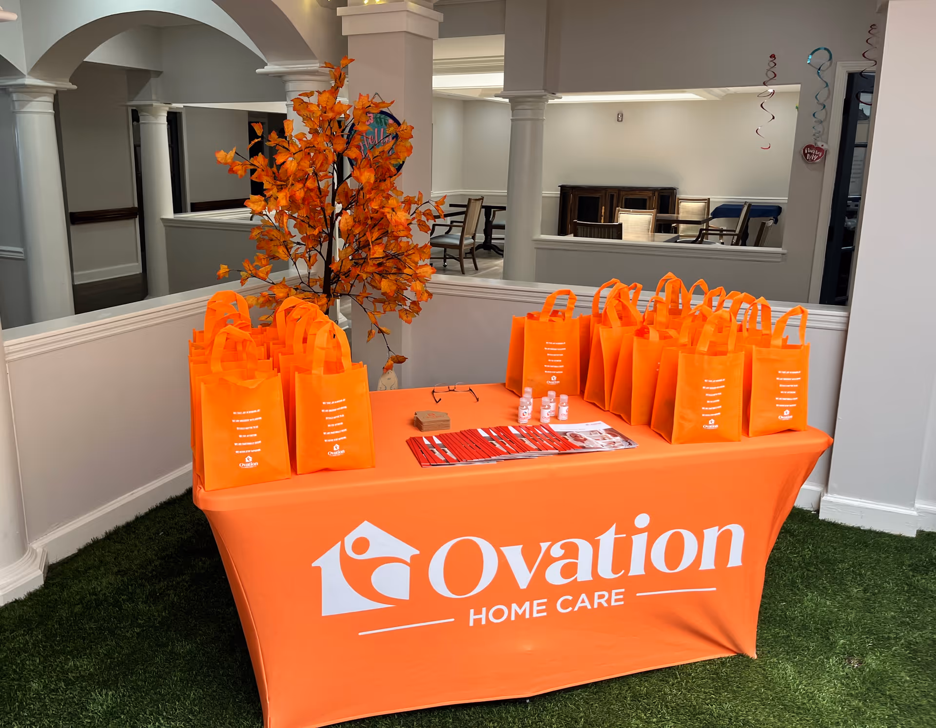 Orange Ovation Home Care bags line a table in a bright indoor atrium.