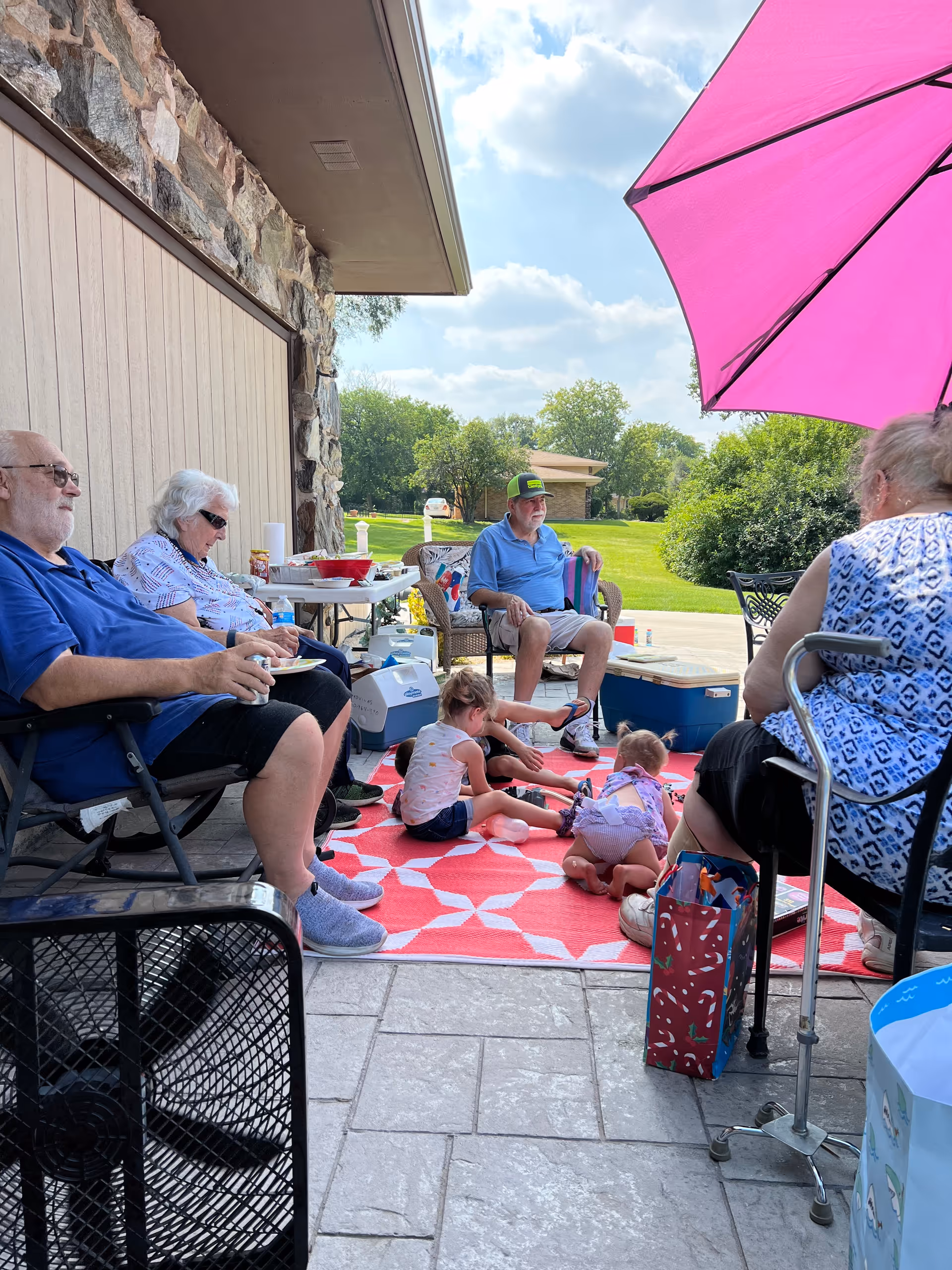 A group of elderly adults and two young children sitting and playing on a red and white patterned outdoor rug under a pink umbrella on a patio next to a stone wall. The adults are seated on chairs around the children, with coolers and snacks nearby. The background shows a green lawn and trees under a partly cloudy sky.