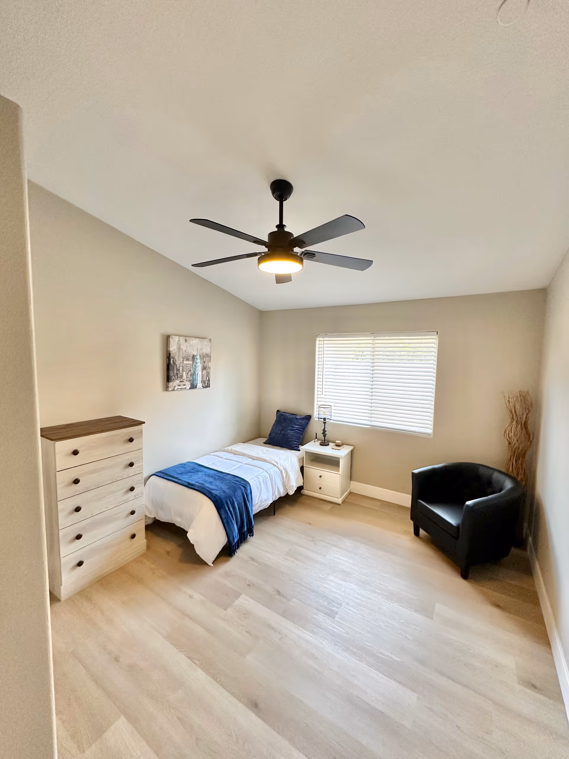 A small, neatly arranged bedroom with a bed, dresser, nightstand, window, and ceiling fan.