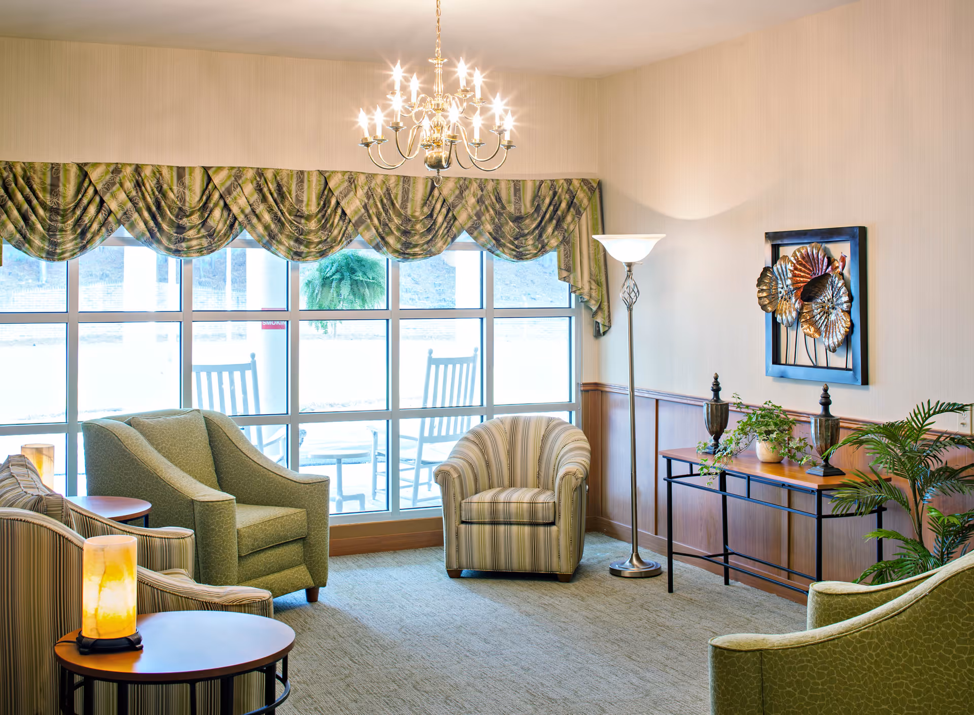 A cozy living room area with large windows covered by green patterned valances. The room features several upholstered armchairs in green and striped fabric, a round wooden side table with a lit lamp, a standing floor lamp, and a console table decorated with plants and metal wall art. Outside the windows, two rocking chairs are visible on a patio.