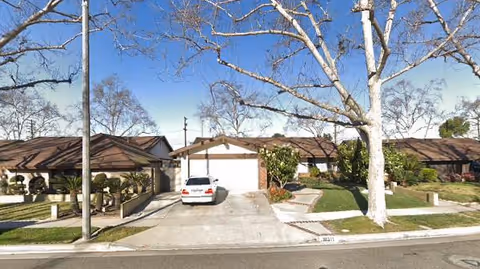 Street view of a residential neighborhood showing single-story houses with garages, a white car parked in a driveway, leafless trees, and a clear blue sky.