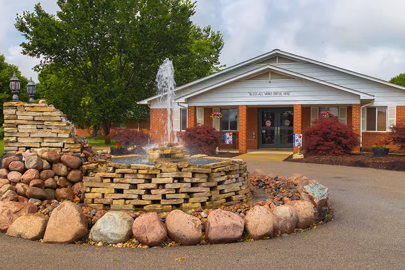 Stone fountain and landscaped driveway in front of the single-story brick entrance to a health and rehab center.
