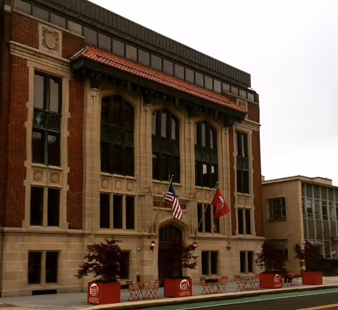 Front facade of a brick building with arched windows, flags, and red planters.