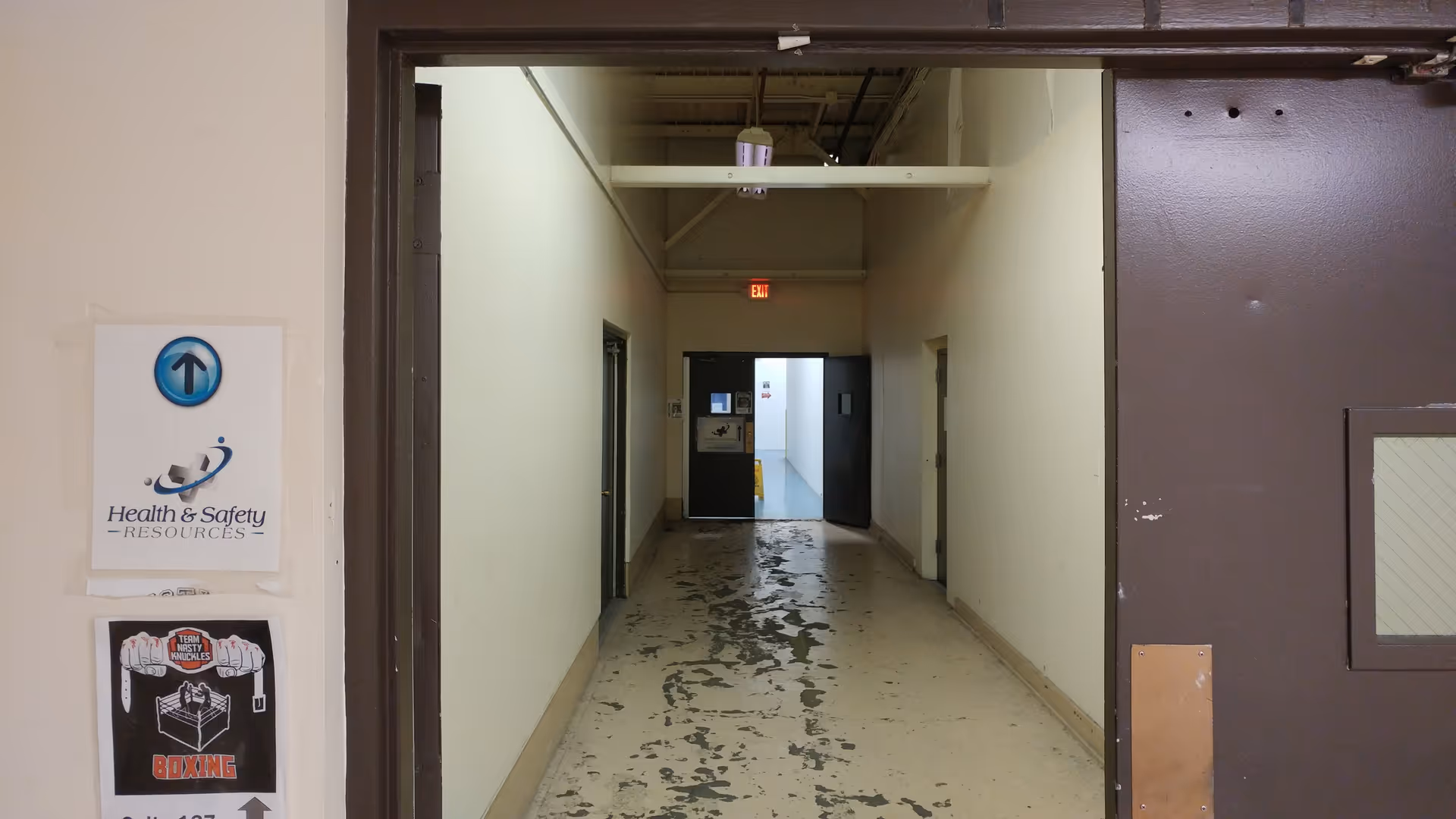 Dim hallway inside a facility with signs, doors, and a peeling floor near the exit.
