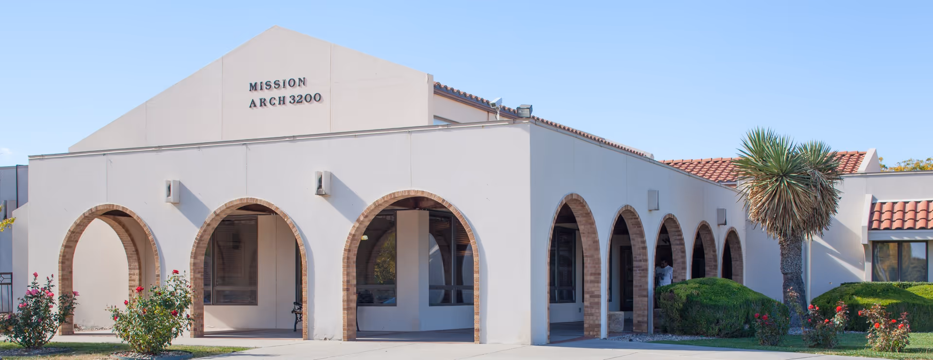 Exterior view of the Mission Arch Center building featuring a white facade with multiple arched doorways and windows, a red-tiled roof, and landscaping including bushes, rose plants, and a palm tree under a clear blue sky.