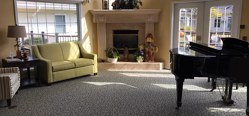 Sunlit common room with a fireplace, green loveseat, grand piano, and large windows.