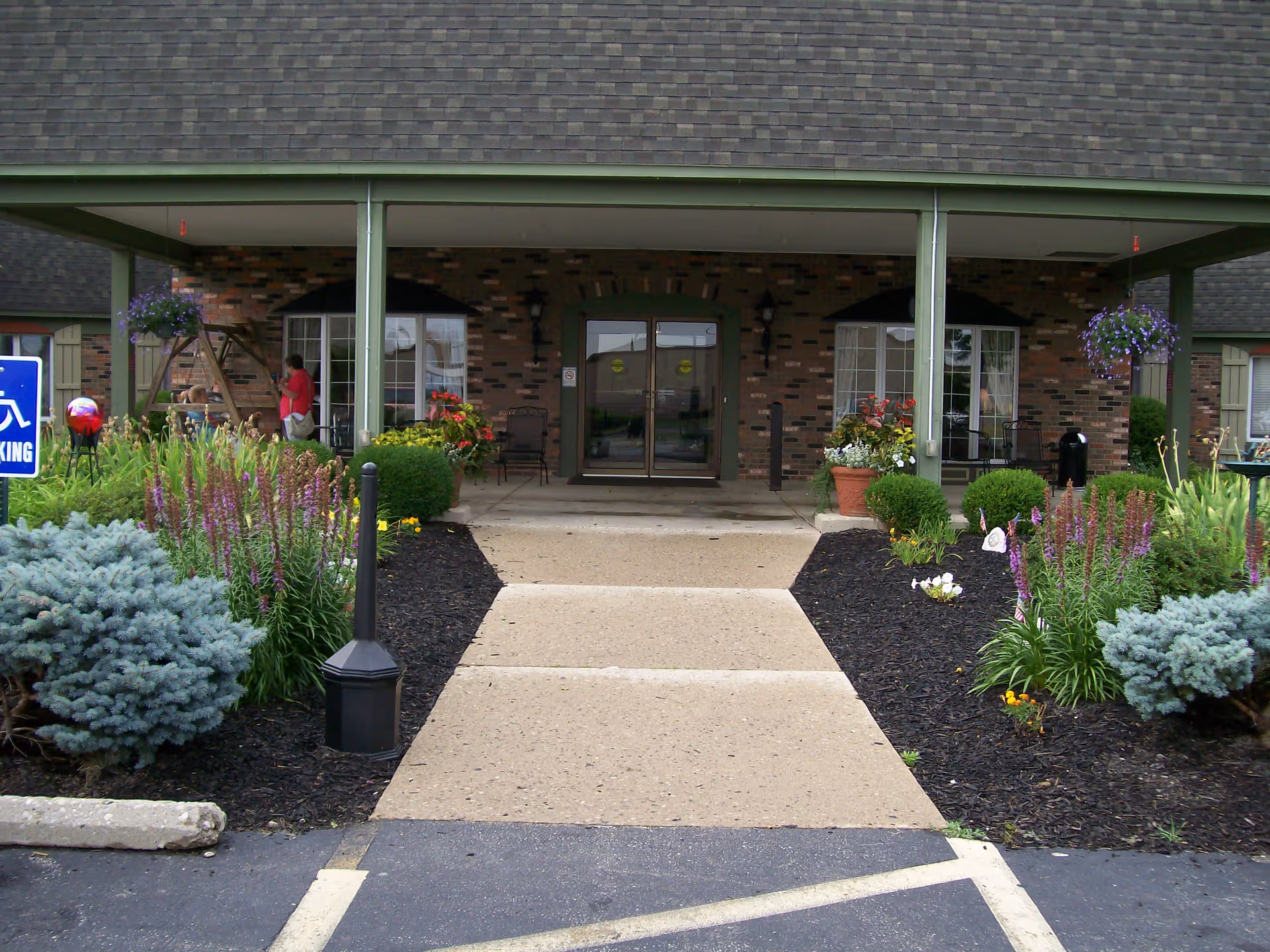 Entrance to a brick building with a covered porch supported by green pillars. There is a concrete walkway leading up to double glass doors. The walkway is flanked by landscaped flower beds with various plants and flowers. A person in a red shirt is visible near the left side of the porch. A blue handicapped parking sign is visible on the left side of the image.