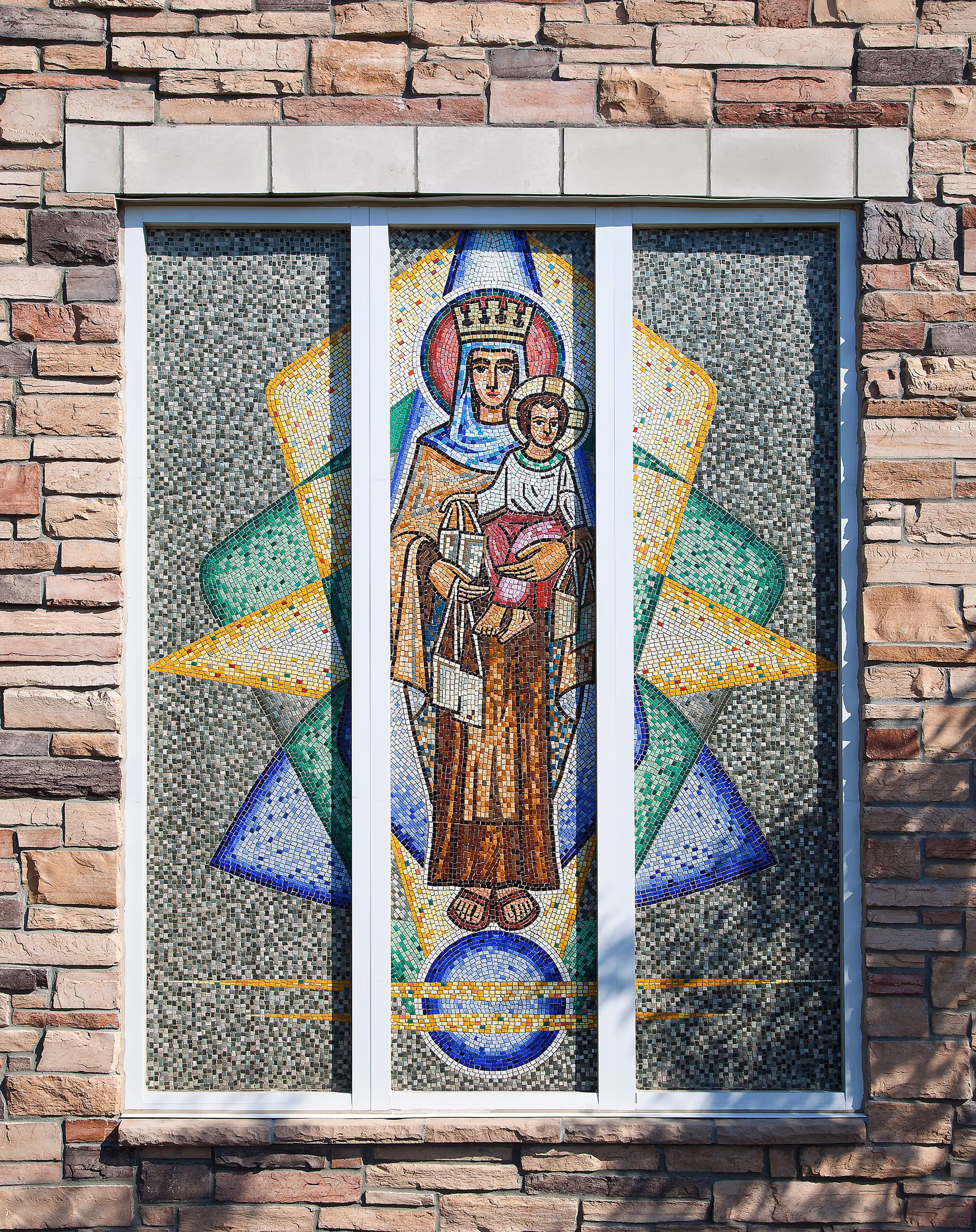 A stained glass window set in a stone wall depicting a religious figure, likely the Virgin Mary, holding baby Jesus. The artwork is composed of colorful mosaic tiles with geometric patterns in the background.