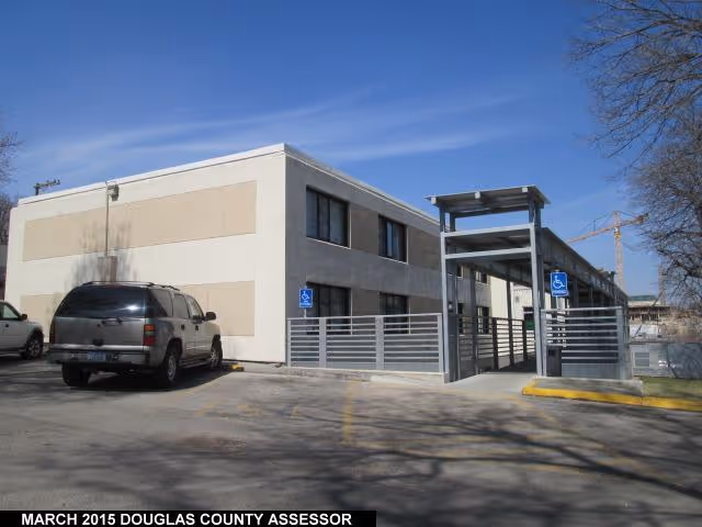 Exterior view of a two-story beige building with a covered entrance and a parking lot with two vehicles, including an SUV. There are handicap parking signs near the entrance. The sky is clear and blue.