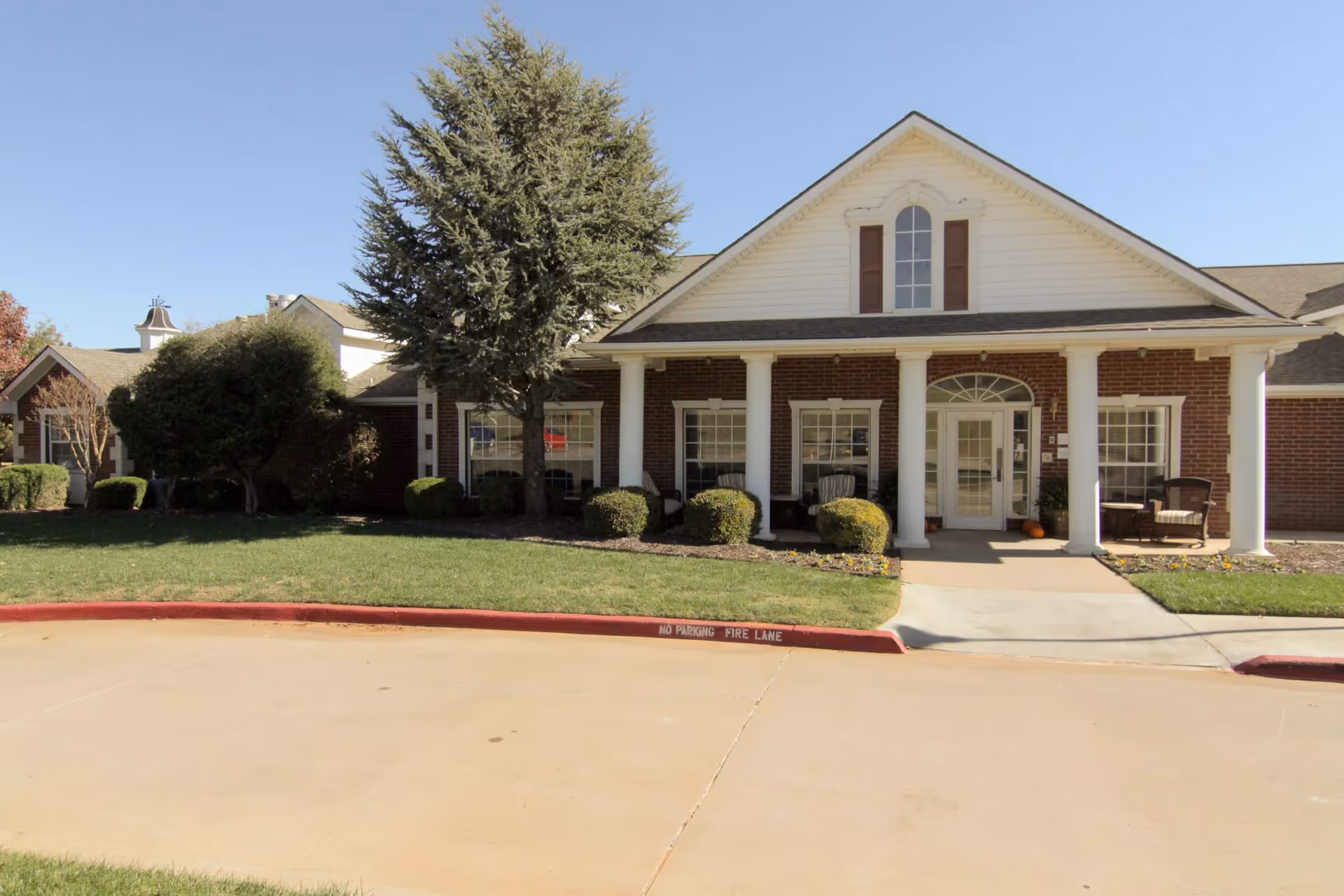 Single-story brick senior living facility entrance with white columns, a small porch and landscaped lawn under a clear sky.