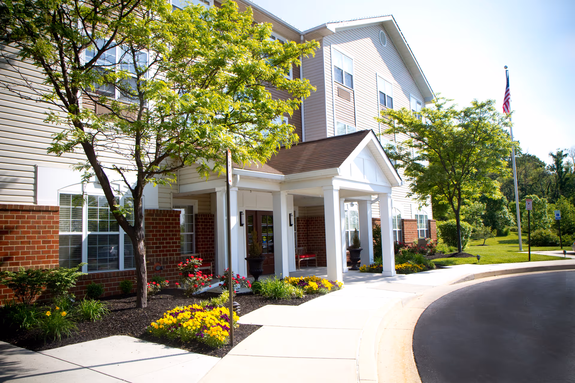 Front entrance of a multi-story senior living building with a covered portico, landscaped flower beds, trees, and a curved driveway.