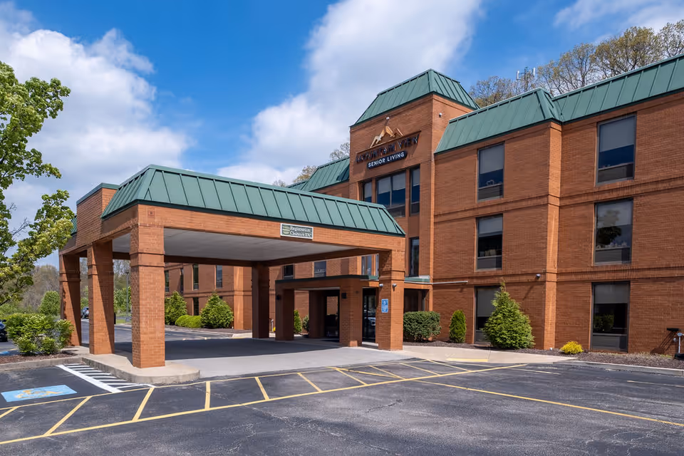 Brick three-story senior living building with a green metal roof and covered porte-cochère entrance under a blue sky.