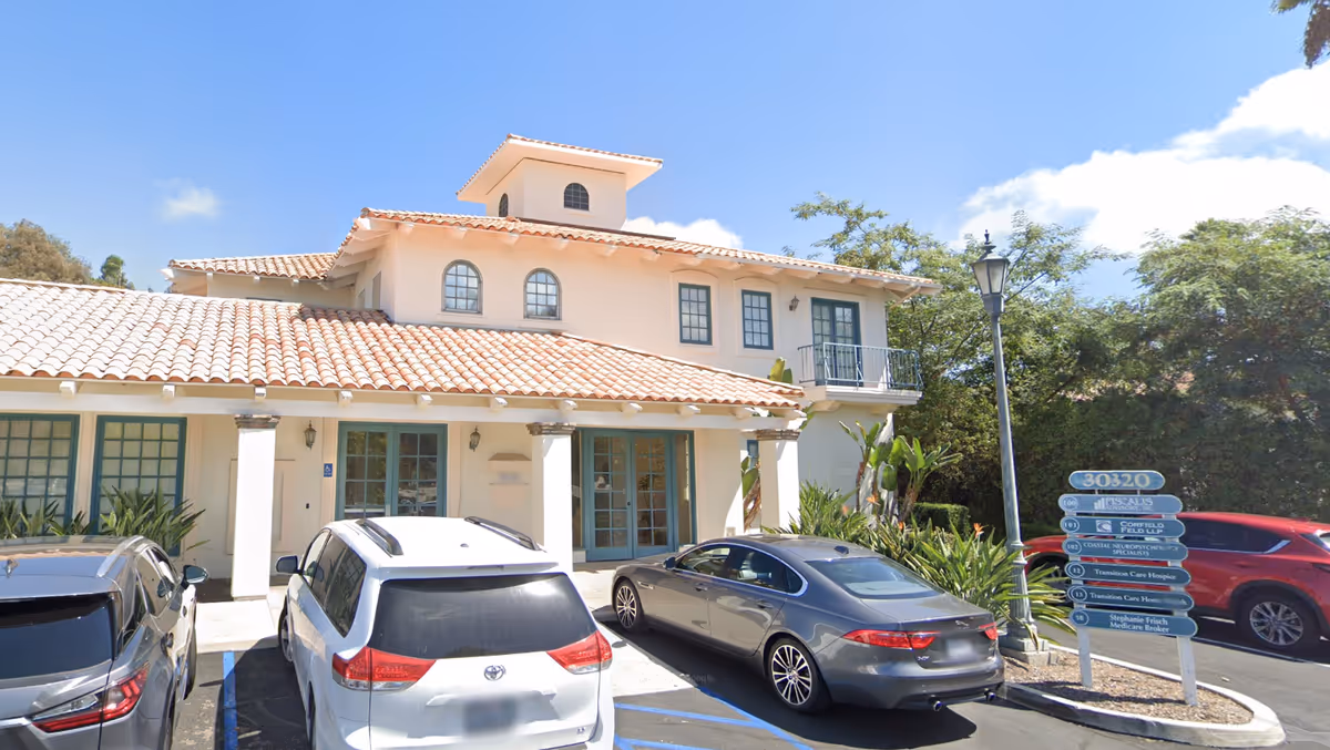 Front exterior of a Spanish-style care facility with parked cars and a signpost.
